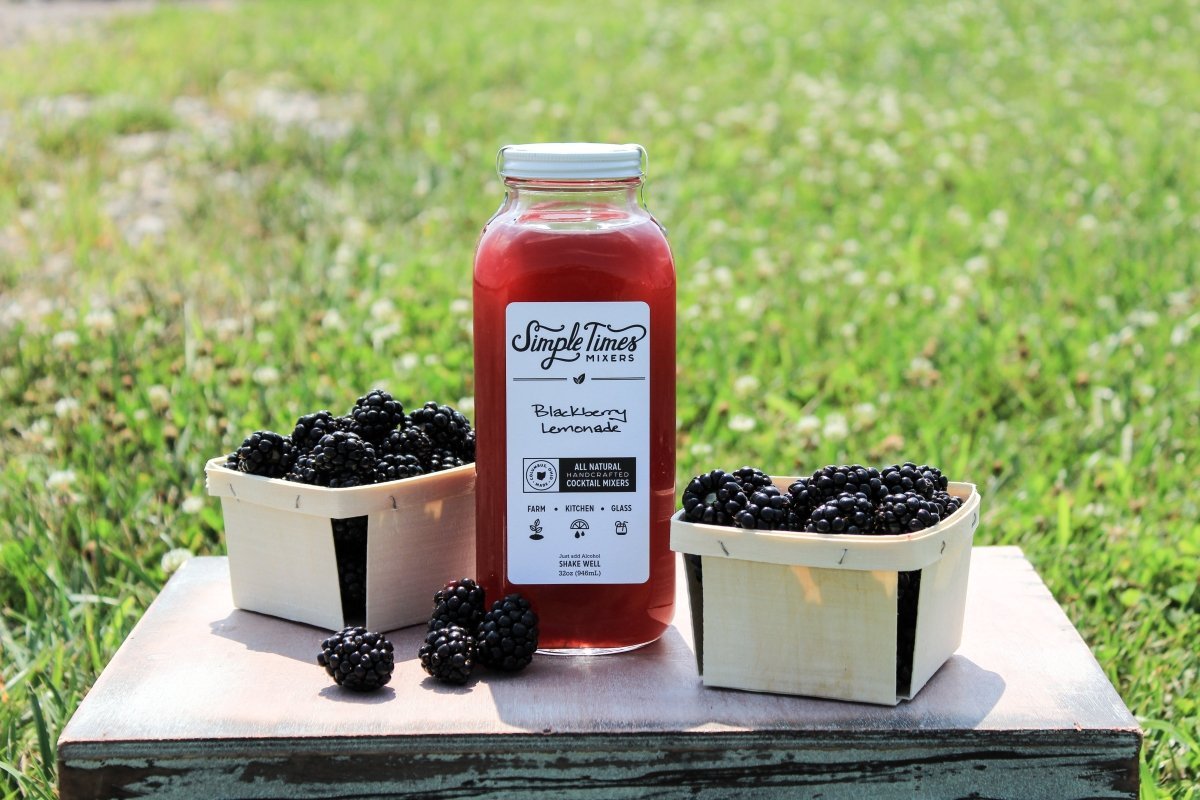 Front view of Blackberry Lemonade mixer bottle showing label text and two baskets of blackberries on a wooden table with a grassy field background
