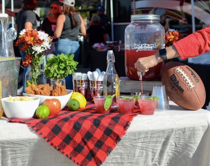 Tailgate party table with red drinks, snacks, fruit, flowers, and a football at an outdoor gathering