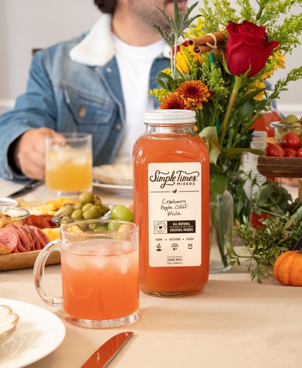 Cranberry apple cider mule mixer displayed at a holiday party with a drink, charcuterie, flowers, and fruit