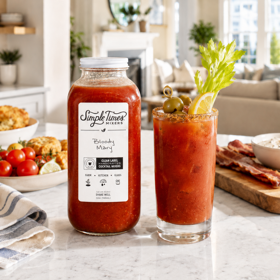 A bottle of Simple Times Farms Bloody Mint sauce stands on a marble countertop beside a tall glass of the prepared drink, garnished with celery, a lemon wedge, and olives, with a blurred kitchen interior in the background.