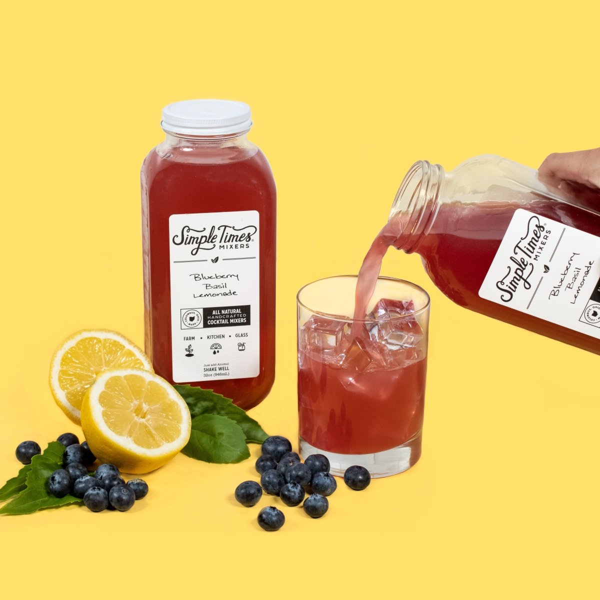 Blueberry Basil Lemonade mixer being poured into an iced glass with lemons, blueberries, and basil nearby