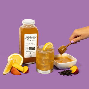 Front of Honey Peach cold-brew tea cocktail mixer bottle and glass with honey drizzle, lemon and peach on purple background