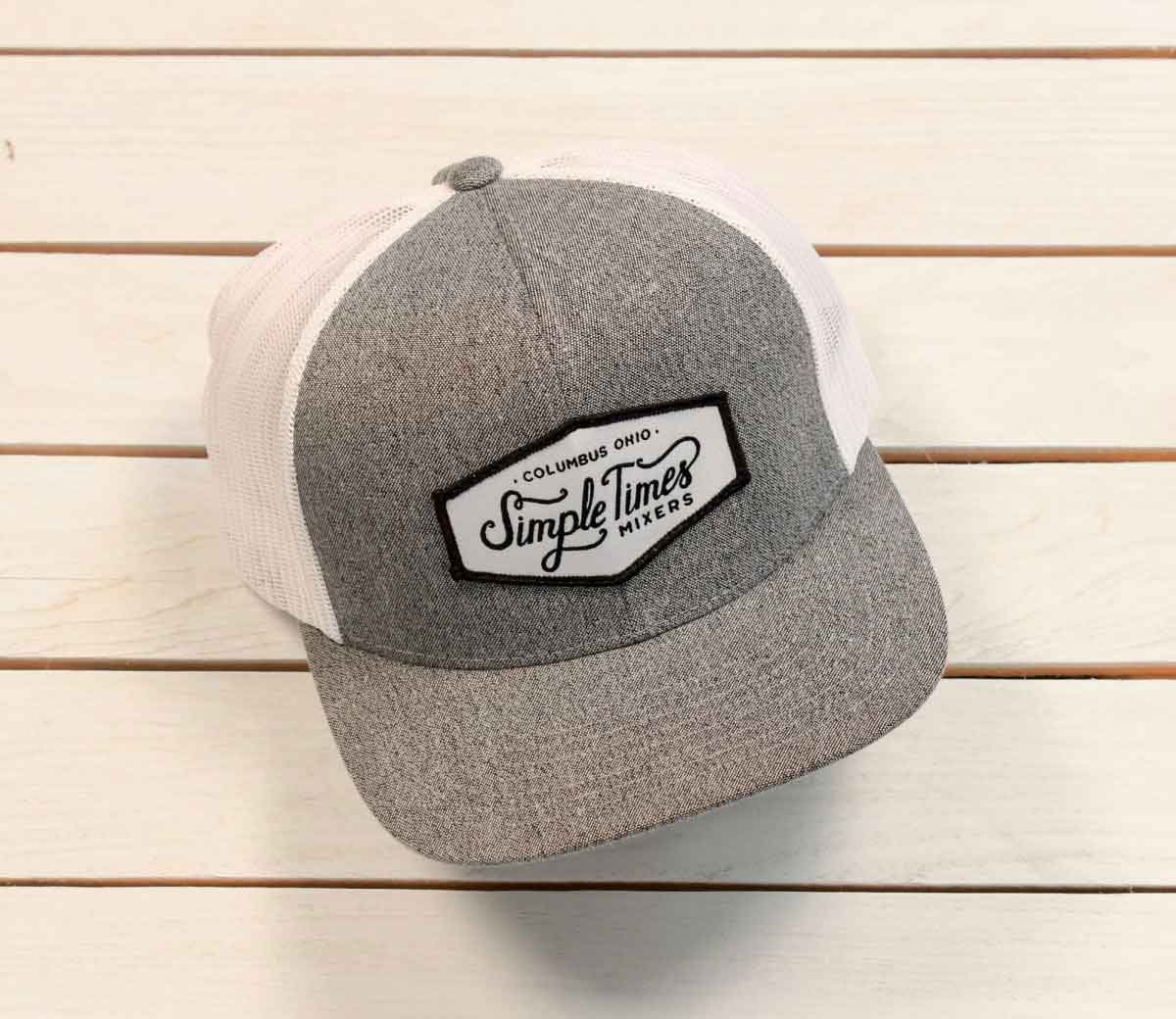 Angled front view of Simple Times Hat showing grey front panel with embroidered Simple Times Mixers patch and white mesh back on light wood slats