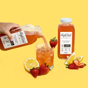 Angled view of Strawberry Lemonade mixer bottle being poured into a glass with strawberries and lemon on yellow background