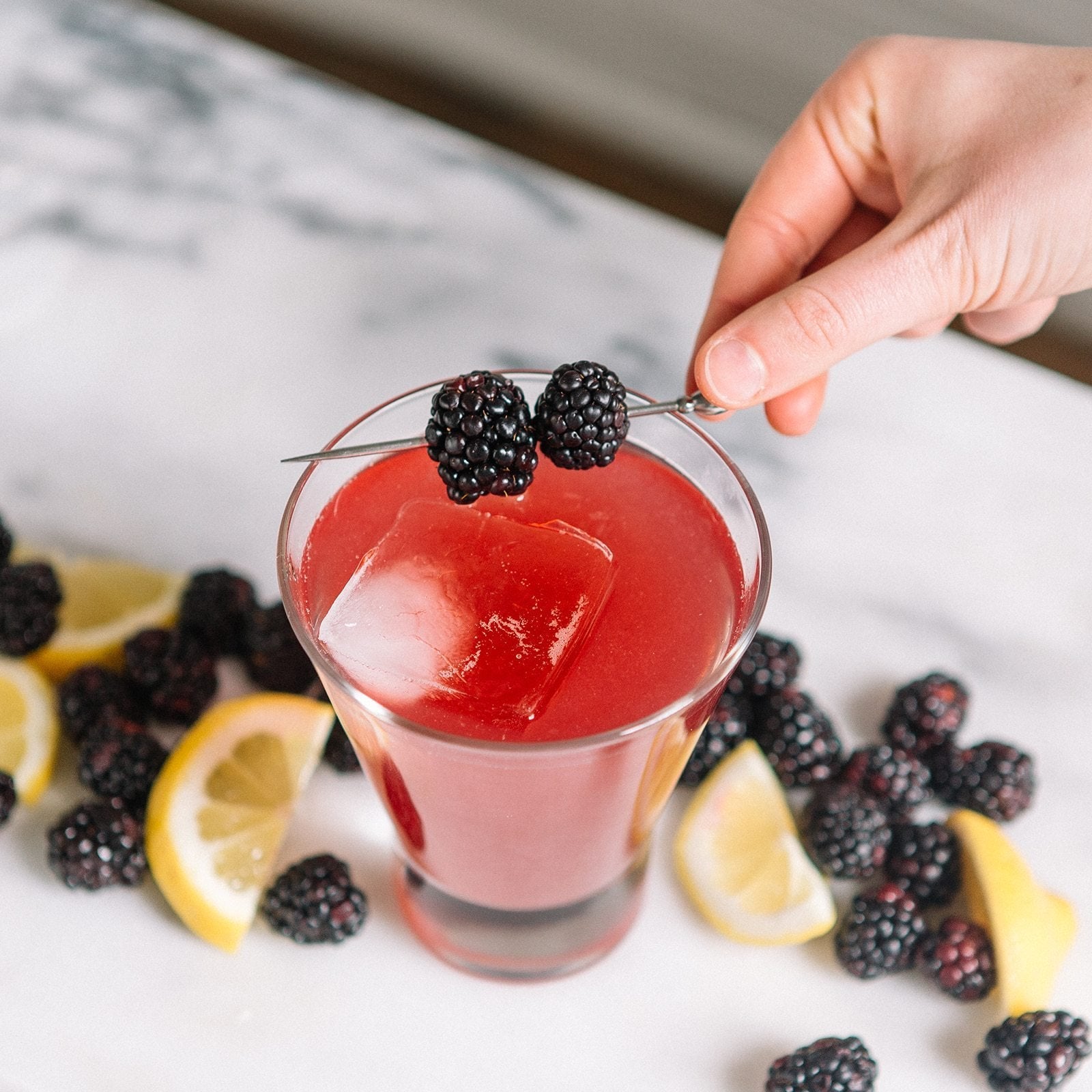 Hand placing a skewer of two blackberries across the rim of a red cocktail glass with ice, surrounded by lemon wedges and blackberries