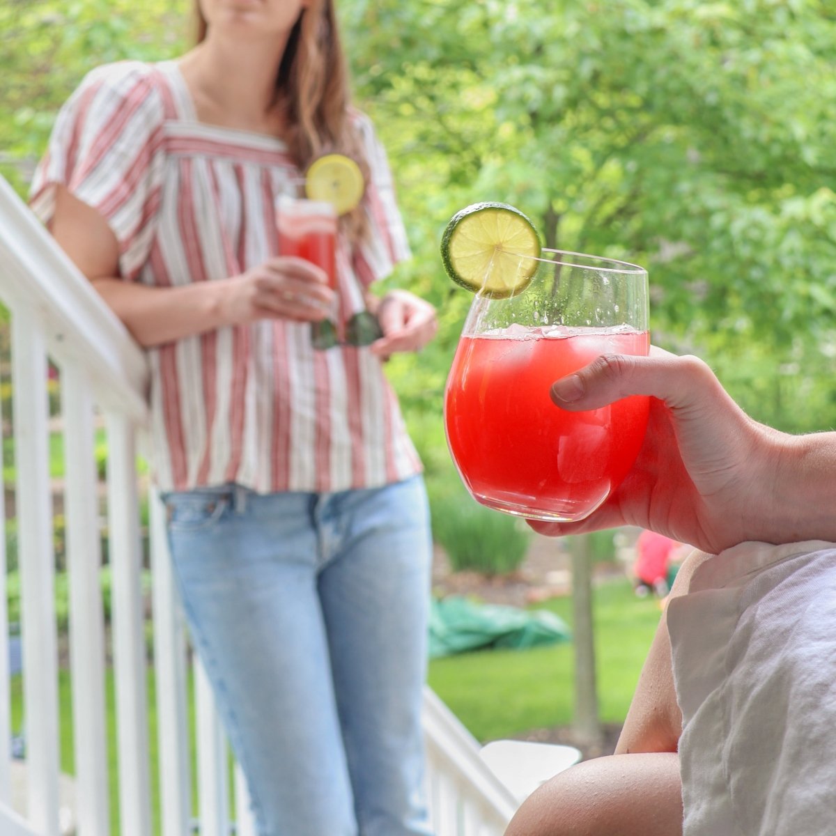 Hand holding a red lemonade cocktail garnished with a lime, another person holding the same drink on a porch with a green backyard