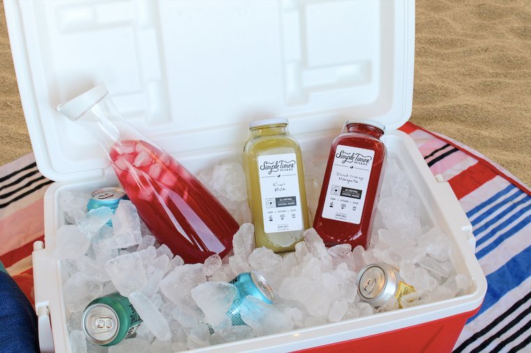 Three Simple Times mixer bottles and several soda cans chilling in a red cooler filled with ice on a striped beach blanket