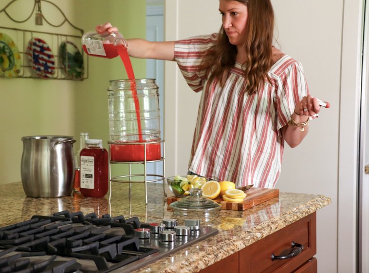Woman pouring red cocktail mixer into a glass dispenser on a kitchen island with sliced lemons and a stovetop