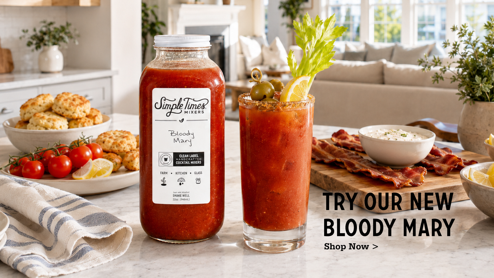 A bottle of Simple Times Mixes Bloody Mary sauce stands next to a prepared Bloody Mary cocktail in a glass, set on a kitchen counter with food props and a living room background. Text on the right reads 'TRY OUR NEW BLOODY MARY Shop Now >'.