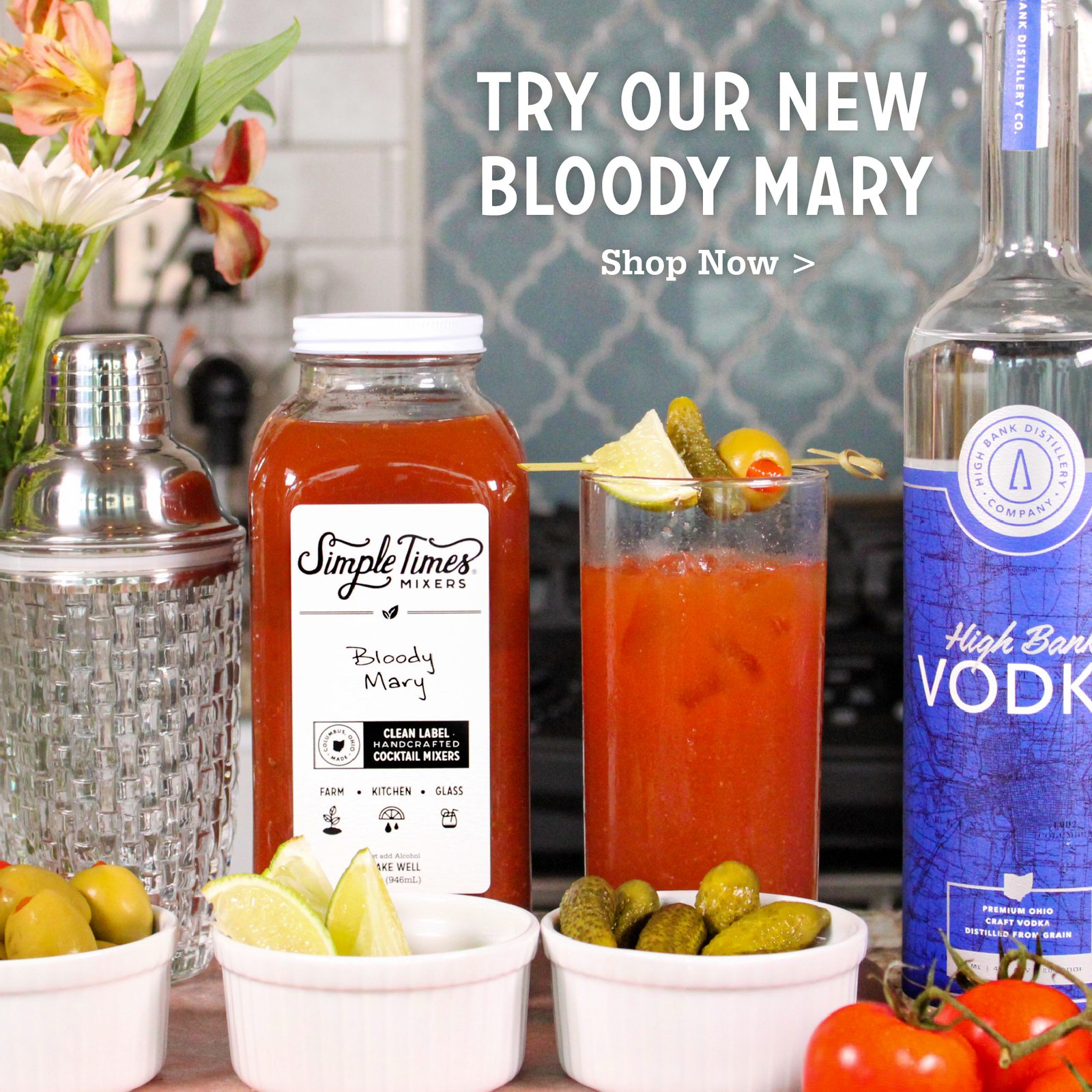 A Bloody Mary cocktail setup with a glass of red drink garnished with pickle slices and lemon, a bottle labeled Simple Times Mixes Bloody Mary, a bottle of high barrel aged vodka, olives, lime wedges, tomatoes, and a cocktail shaker on a kitchen counter. Text overlay reads 'TRY OUR NEW BLOODY MARY Shop Now >'.